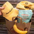 Two banana bread baddie classic caps, a bag of flour, and a banana on a wooden surface.