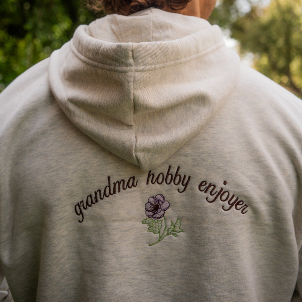 Person wearing a oatmeal banana bread baddie branded hoodie with 'grandma hobby enjoyer' text and flower design outdoors.