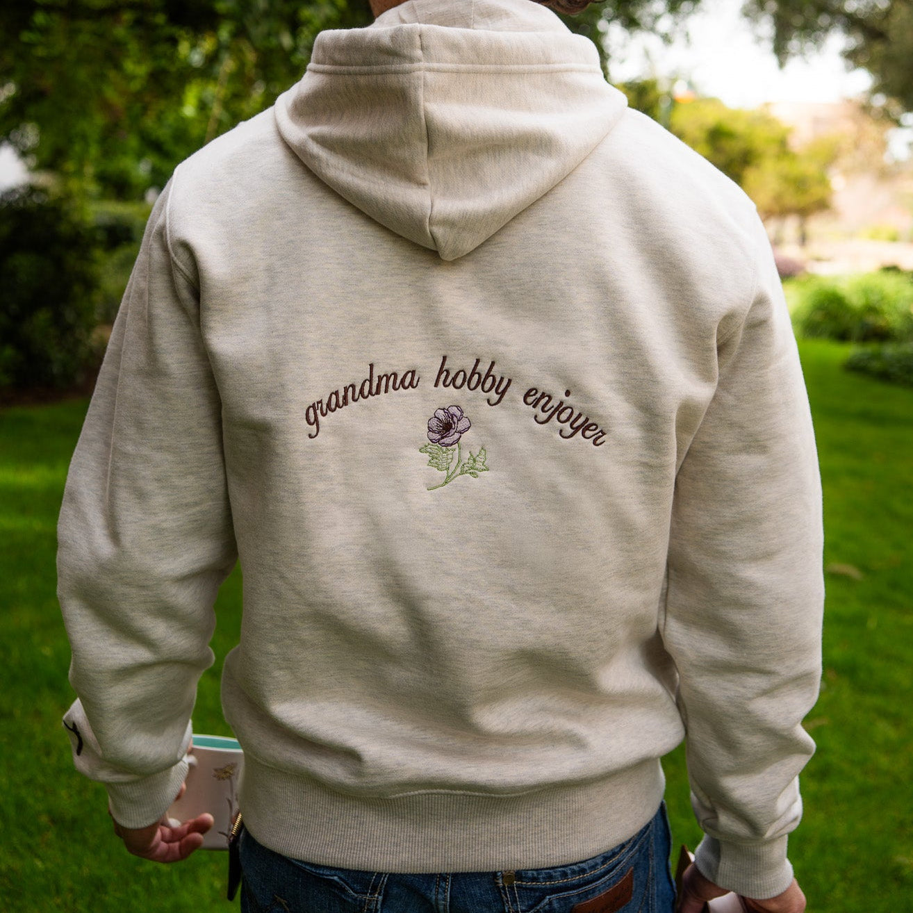 Person wearing a hoodie with 'grandma hobby enjoyer' text and a lavender flower graphic outdoors.