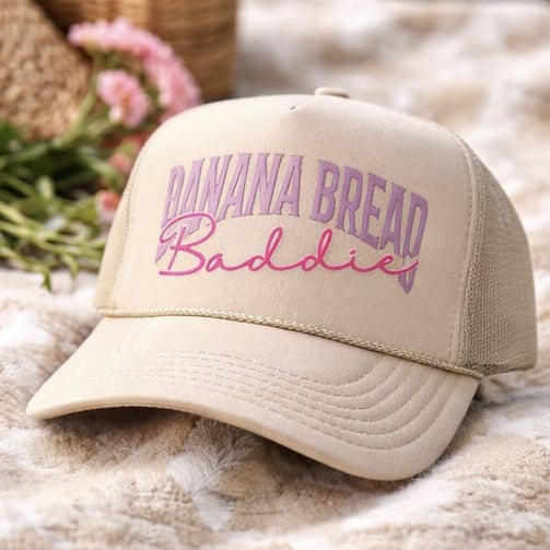 Beige cap with 'Banana Bread Baddie' text on a textured surface with flowers and wicker basket in the background