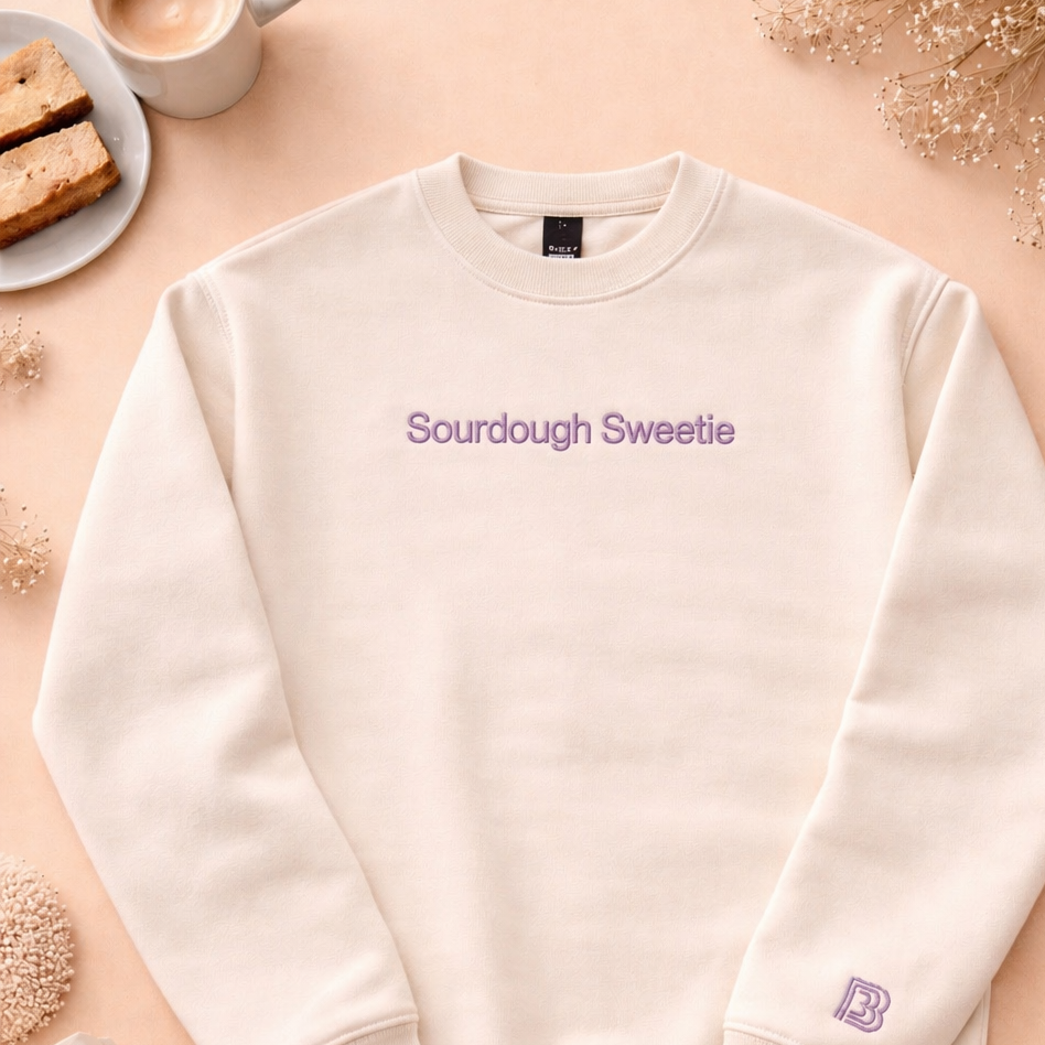 White sweatshirt with 'Sourdough Sweetie' text on a beige surface with a cup of coffee and snacks.