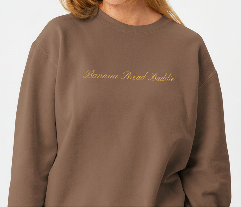 Woman wearing a brown sweatshirt with 'BANANA BREAD BADDIE' text on a white background in script font